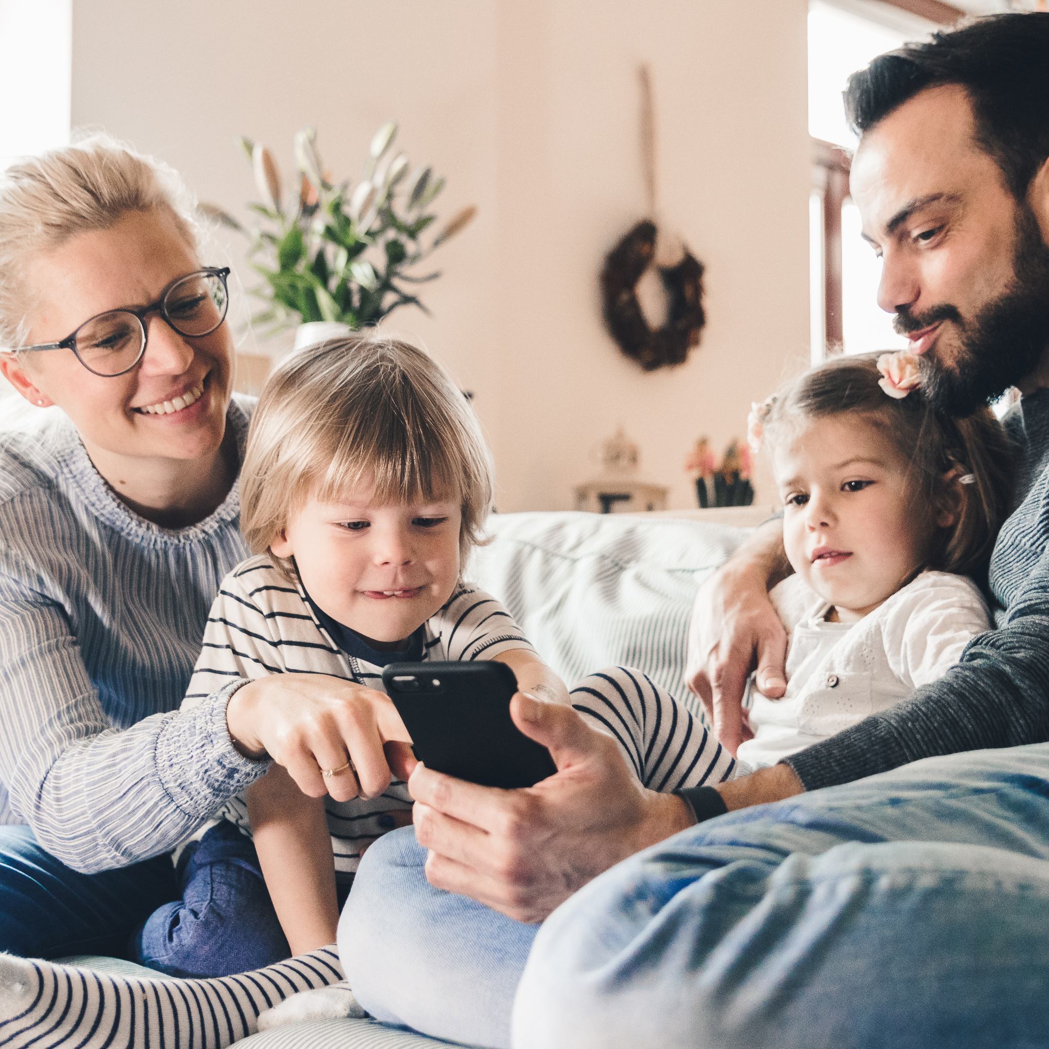 Familie sitzt auf der Couch und schaut gemeinsam auf ein Smartphone. 